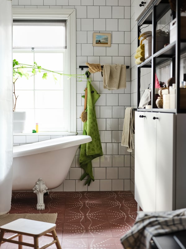 A white-tiled bathroom with plants in the window, a bath behind a shower curtain and an ENHET wall storage combination.