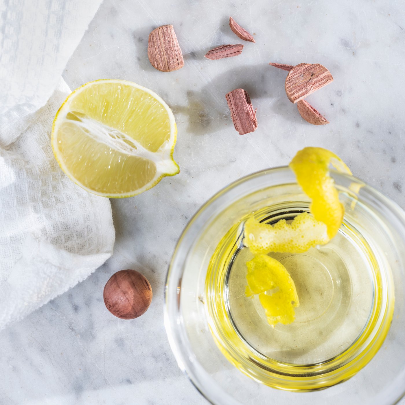 Sklenice s čirou tekutinou a kůrou citronu na mramorovém povrchu, vedle půlka citronu, dřevěné kousky a bílé utěrky – svěží dekorace inspirovaná IKEA.