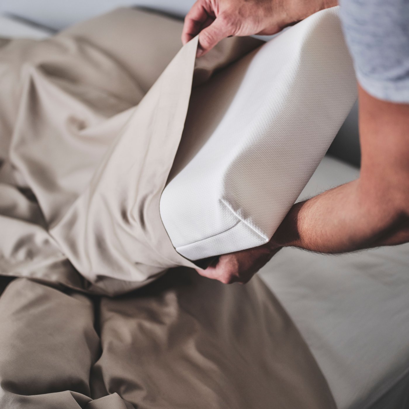 A person is putting a white PAPEGOJBUSKE ergonomic pillow for side/back sleeper into a beige satin pillowcase.