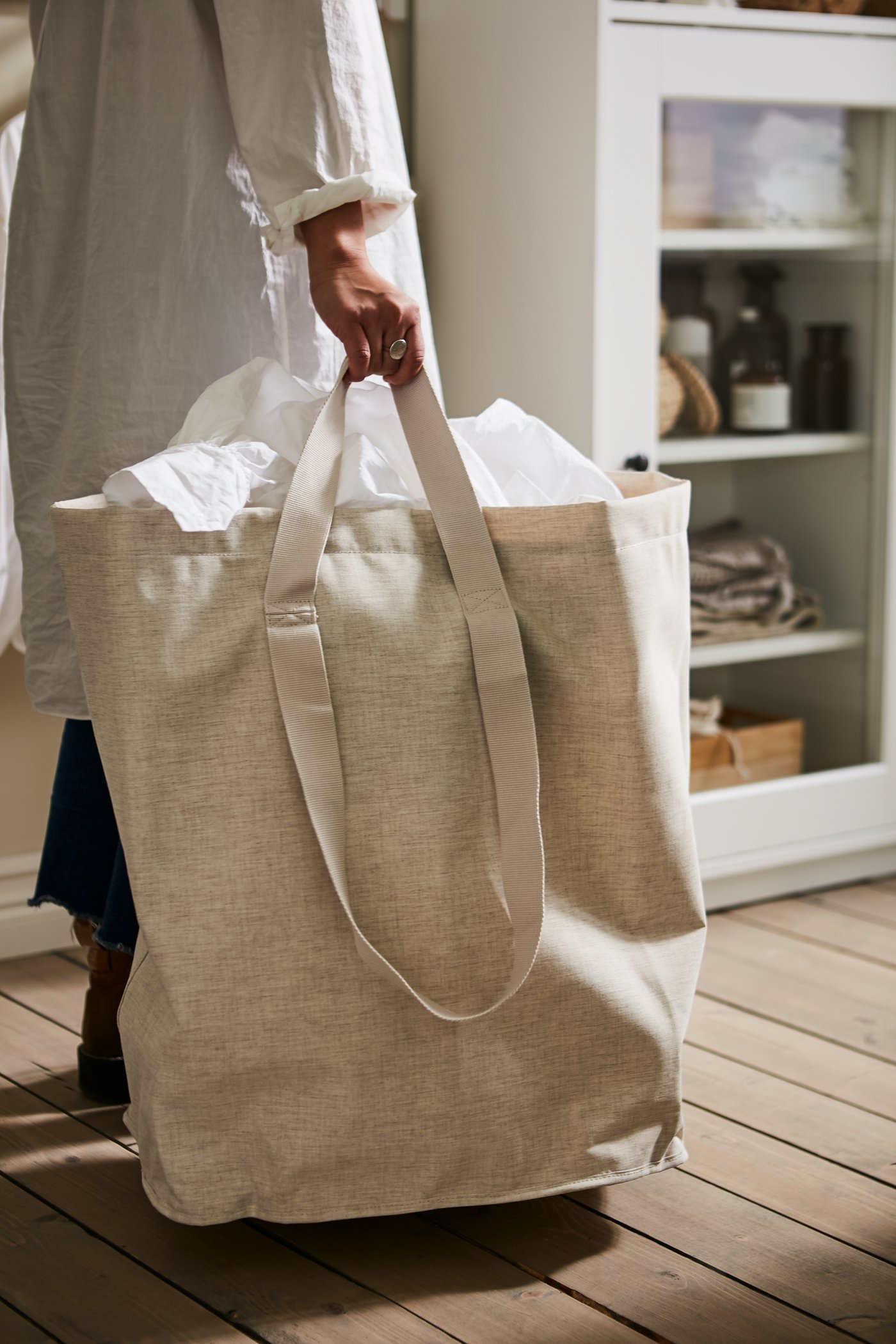 Una persona lleva una bolsa para ropa sucia PURRPINGLA color beige, llena de sábanas blancas, en un hogar con piso de madera.