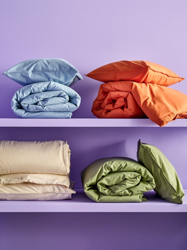 Four ÄNGSLILJA duvet covers and pillowcases in different colours are folded and placed on two lilac shelves.