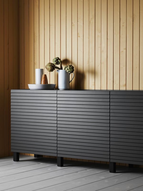 Nahaufnahme eines Sideboards mit Türen aus quer verlaufenden Holzlamellen steht vor einer Holzwand aus längs verlaufenden Paneelen. 