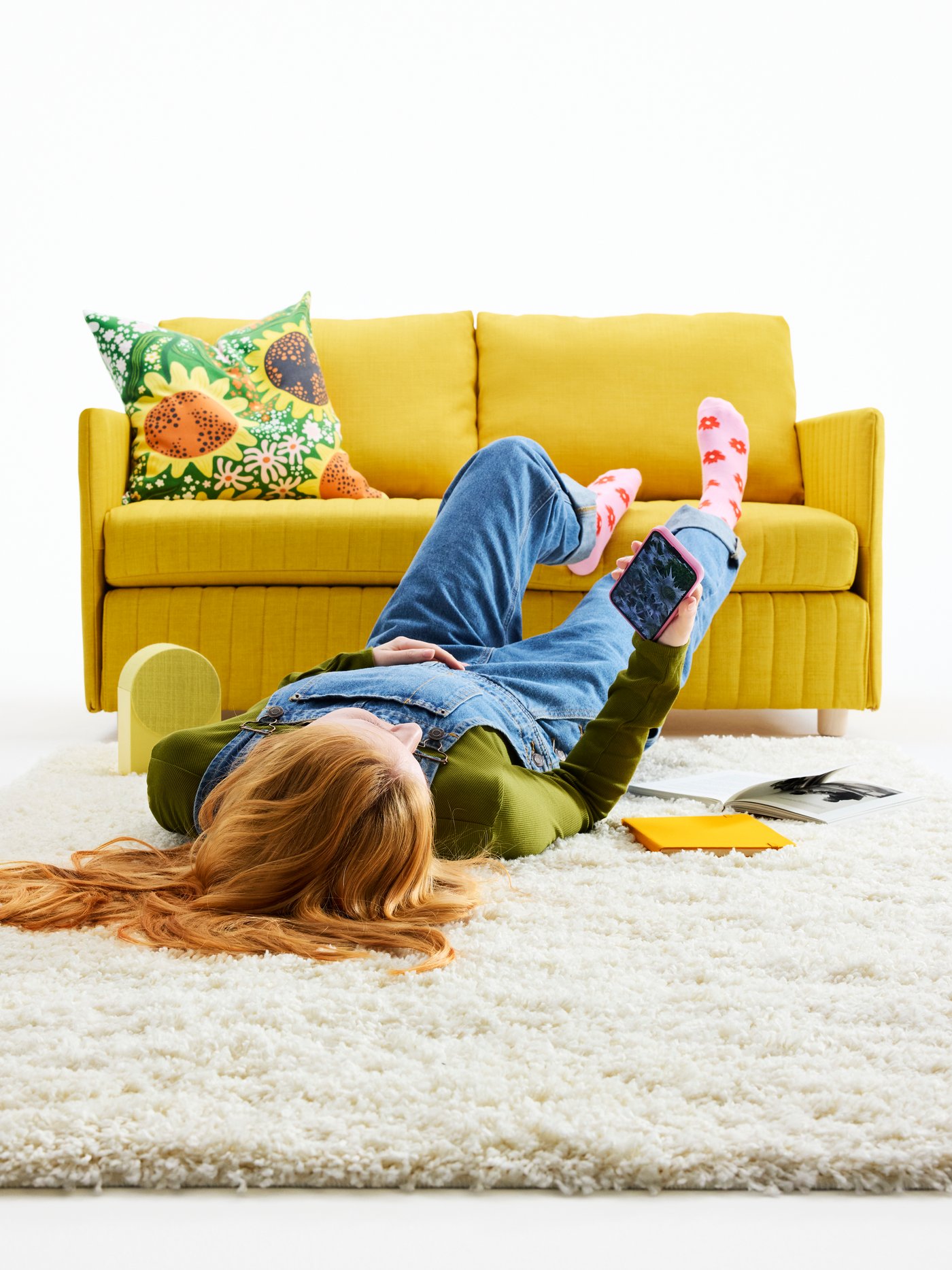 Eine Person liegt auf einem weichen ÄRENDE Teppich vor einem gelben Sofa. Bunte Akzente schaffen eine lebendige Atmosphäre.