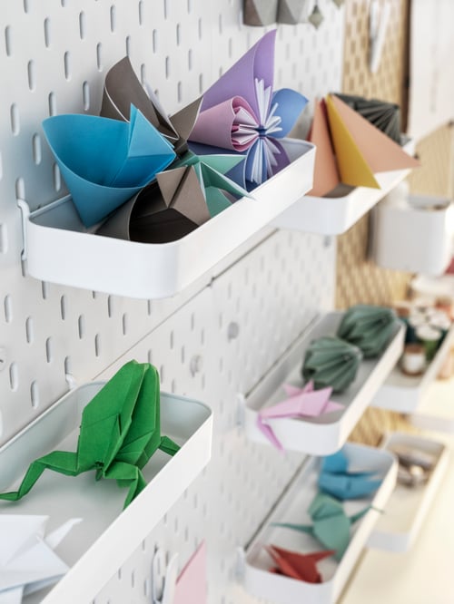A close-up of many white SKÅDIS shelves attached to matching, white pegboards. The shelves hold different origami creations.