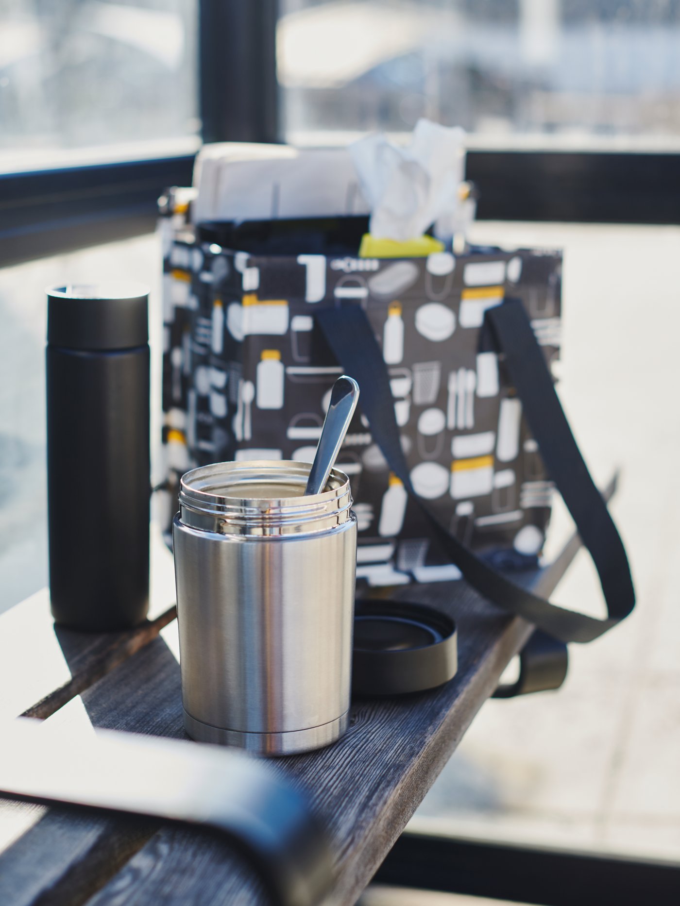 An open EFTERFRÅGAD food vacuum flask with a spoon. Placed in front of a FLADDRIG lunch bag on a sunlit public bench.