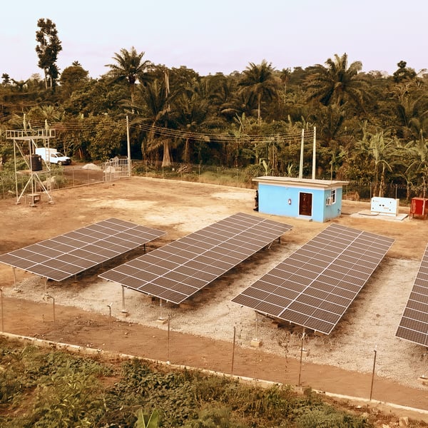 An aerial view of a solar power installation with several rows of large solar panels arranged neatly on a cleared patch of land. In the background, there is lush greenery with numerous trees, and a small blue building with a white roof is situated near the solar panels. 