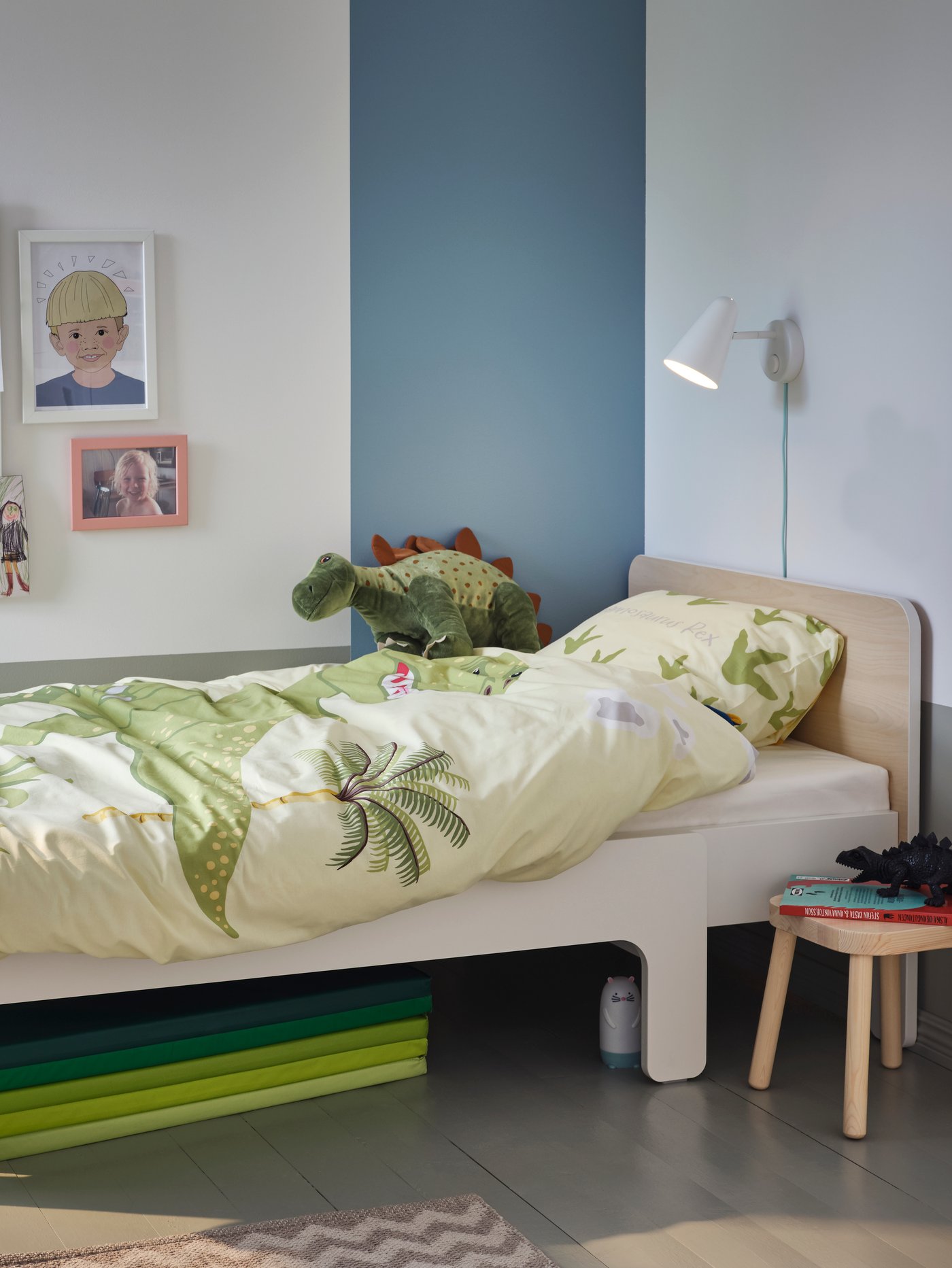 A SLÄKT extendable bed in white/birch, with JÄTTELIK bedding and a soft toy dinosaur on top, stands in a corner.