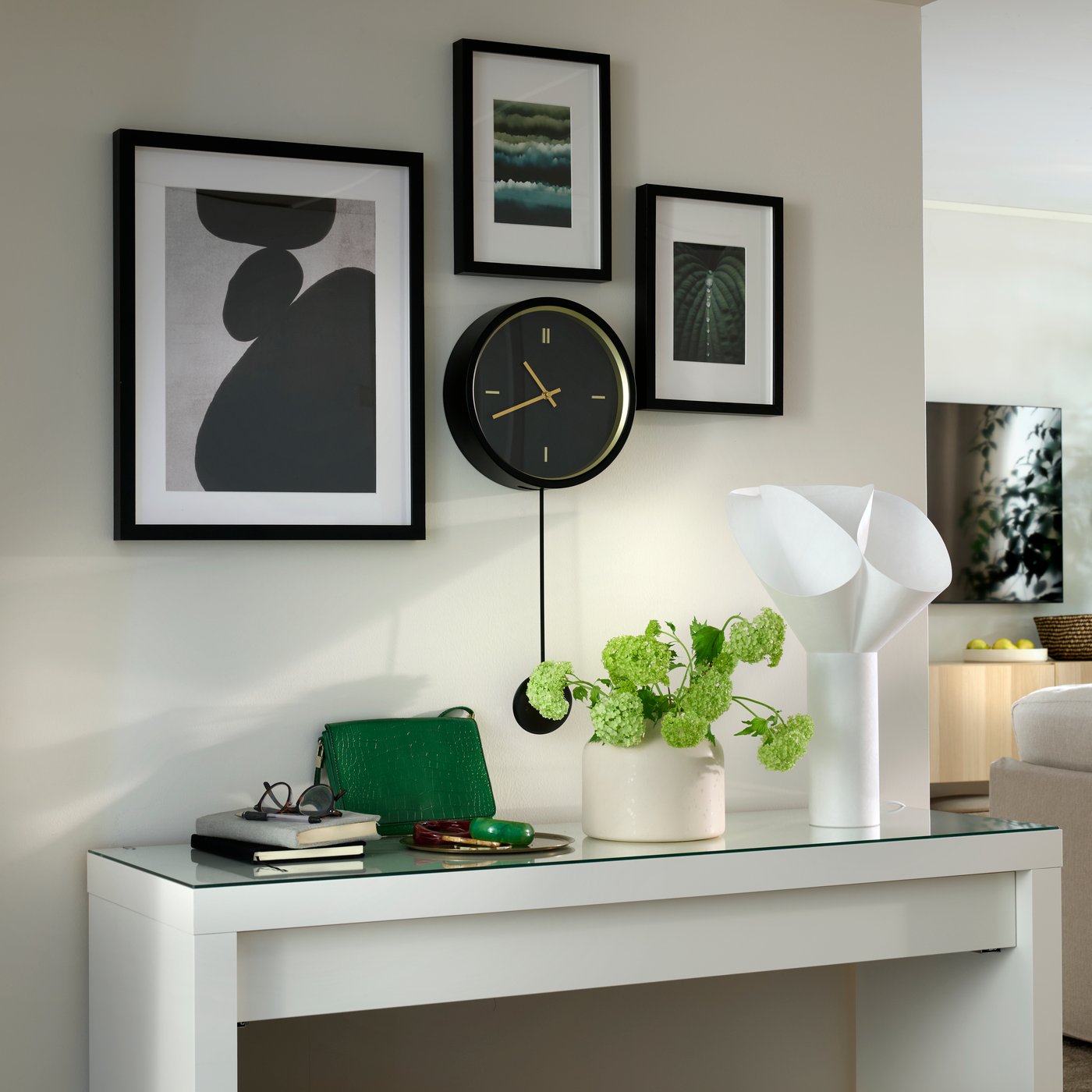 Œuvres d’art encadrées autour d’une horloge murale STURSK noire, le tout accroché au-dessus d’une console sur laquelle est posée une lampe de table BÄCKNATE blanche.