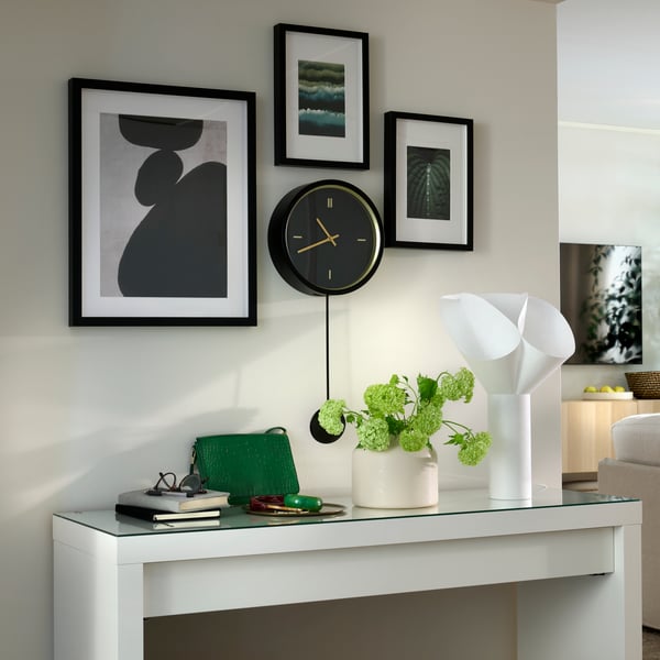 Framed art hanging around a black STURSK wall clock, all placed above a side table with a white BÄCKNATE table lamp on it.