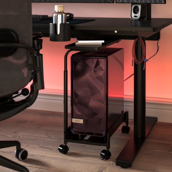 A black LÅNESPELARE computer cart with a shelf is set under a MÅLOMRÅDE gaming desk. Pinkish light glows behind the desk.