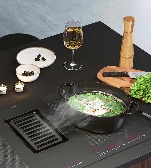 Ein FÖRDELAKTIG Induktionskochfeld mit integrierter Dunstabzugshaube, auf dem ein Topf steht. Daneben auf der Arbeitsplatte steht ein Glas Wein, eine Pfeffermühle, ein Schneidebrett mit einem Messer und Kräutern und 2 Teller.