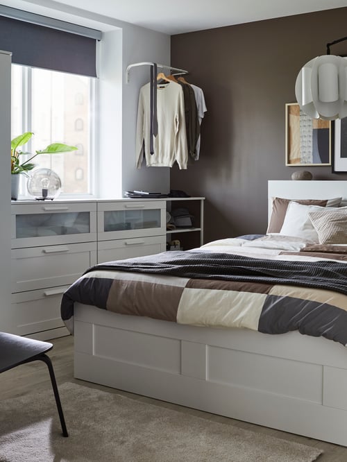 Bedroom with bed frame, storage drawers, headboard, storage units, duvet cover and pillowcases, clothes rack, table lamp, and rug.