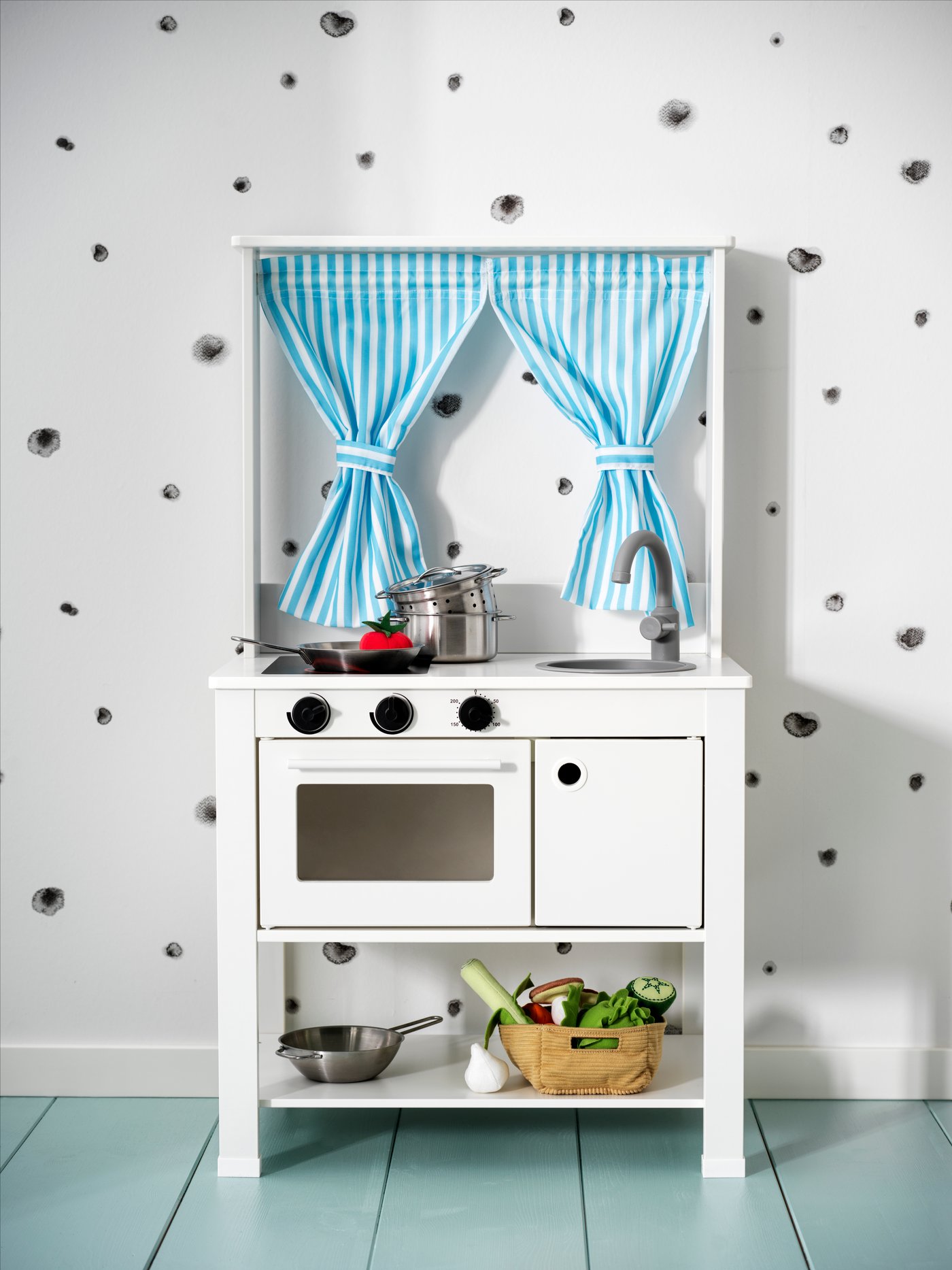 Una cocina de madera SPICIG blanca apoyada en una pared blanca con puntoos de colores. La cocina tiene cortinas de tela celestes con blanco.