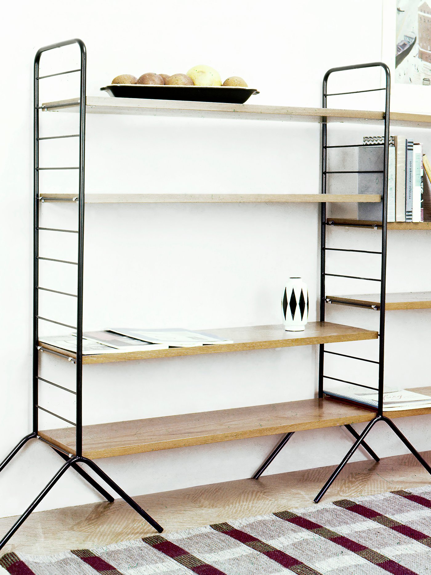 Es ist das IKEA HANSA Regal von 1957 zu sehen, das aus dunklen Metallseitenteilen und Beinen und Holzböden besteht.