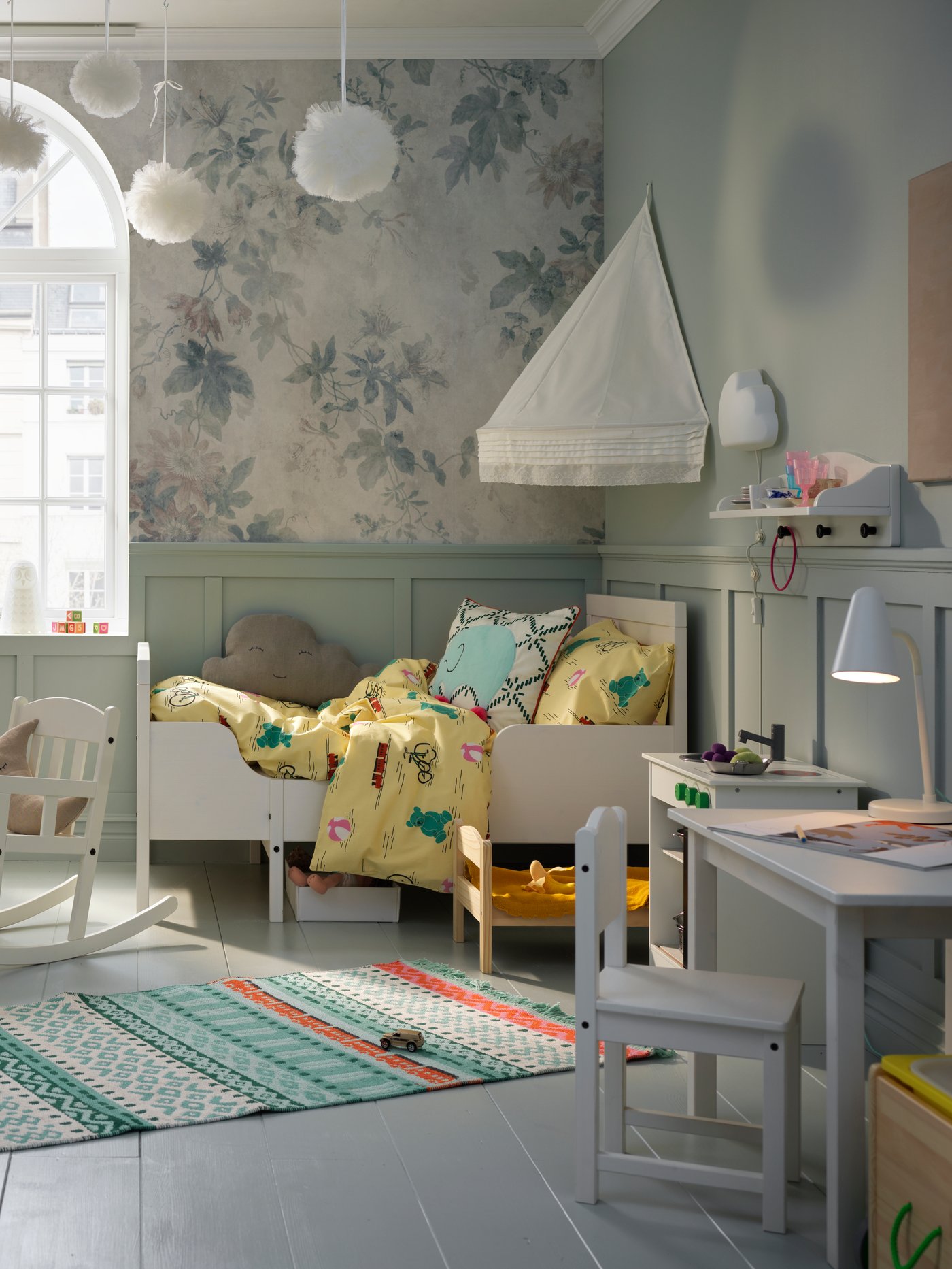 Dormitorio infantil con una cama extensible blanca, una funda nórdica, un dosel, una mecedora, una cocina de juguete y una alfombra.