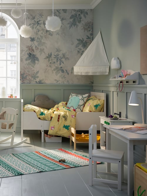 A children's room featuring a white extendable bed, a quilt cover, a bed canopy a rocking chair, a play kitchen and a rug.
