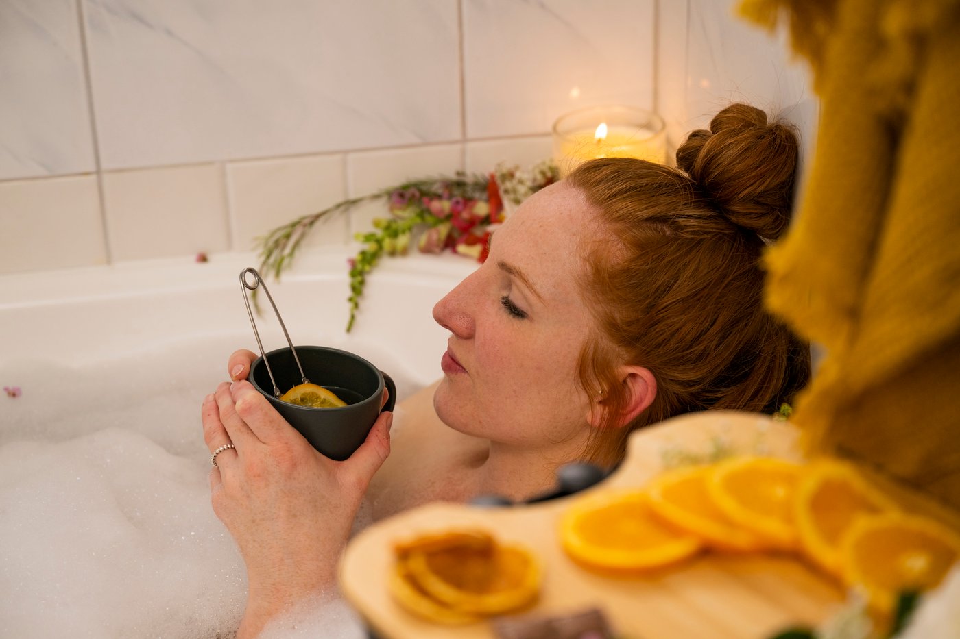 Woman lying in a bubble bath surrounded by candles holding a warm cup of tea with sliced oranges placed on a trolley next to the tub.