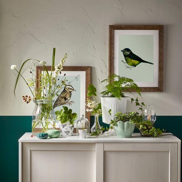 Un mobile bianco con diverse decorazioni: ci sono cornici con stampe di uccelli, vasi dai colori tenui con piante BEGÄRLIG e VILJESTARK, una lanterna e scatole conteinori sui toni del verde.