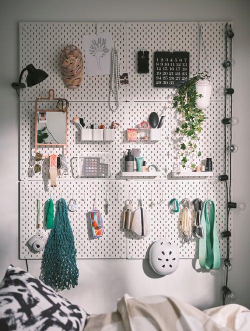 White pegboards mounted on a wall containing a magazine holder, a LED lighting chain, a mirror, a plant, and other items.