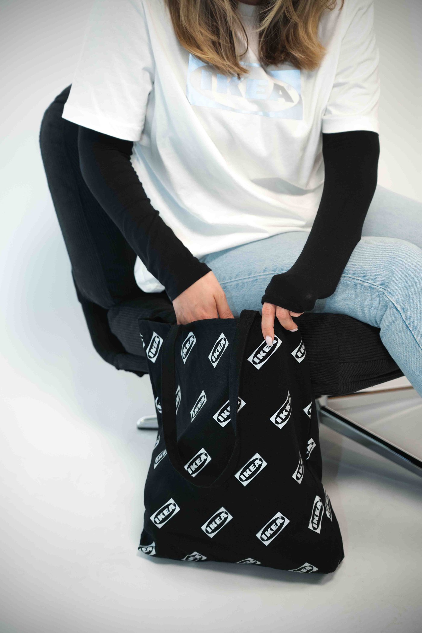Person sitter på stol i vit t-shirt och blå jeans, sträcker sig ner i svart IKEA tygpåse.