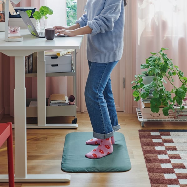 En person står på en grågrön FREIVID ståmatta vid ett vitt skrivbord sitt/stå och skriver på en bärbar dator.