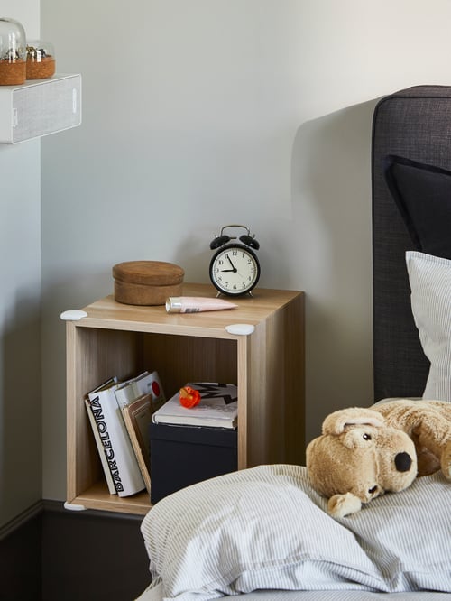 Une étagère murale aspect bois est fixée sur un mur près d’un lit avec des protections d’angle UNDVIKA triangulaires en plastique blanc à chaque angle.