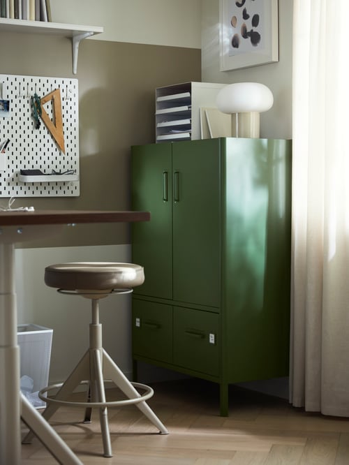 A dark green IDÅSEN cabinet with drawers and doors in the corner of a room with a table lamp and an organiser on top.