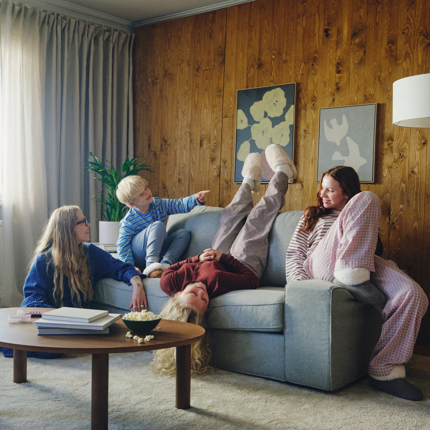 KIVIK-kaksipaikkainen Tibbleby beige/harmaa -sohva on sijoitettu puupaneeliseinää vasten. Sohvalla rentoutuu yksi aikuinen ja kolme lasta yhdessä.