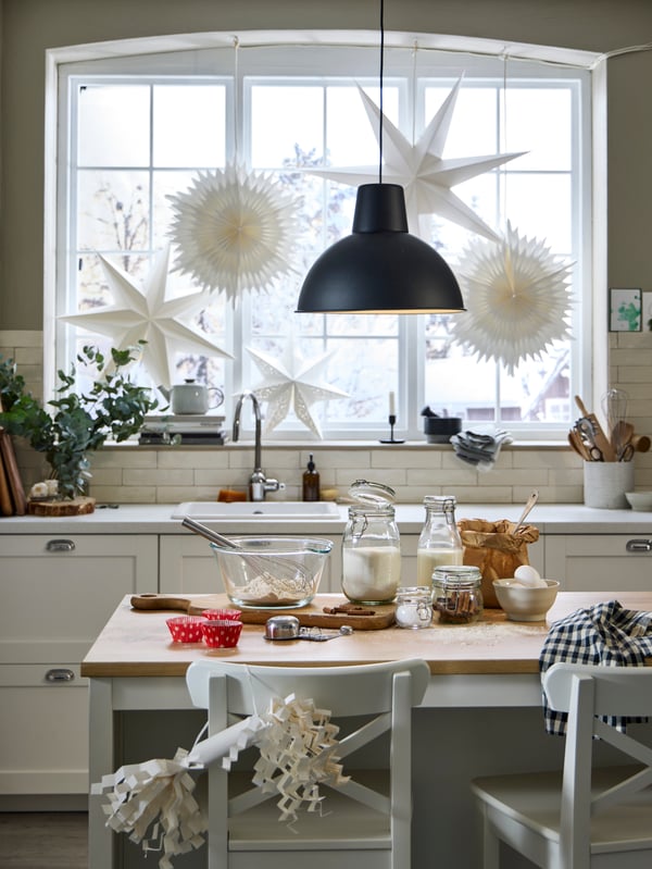 Un cucina bianca con gli ingredienti per un dolce sul tavolo e lampade con decorazioni appese alla finestra. 
