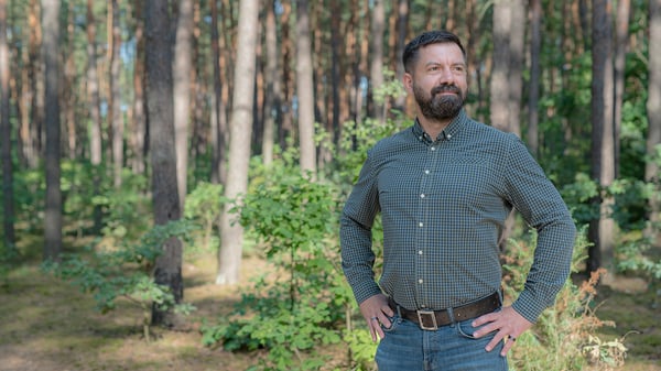Maciej è in un bosco illuminato dal sole, con una maglietta a quadri e dei jeans.