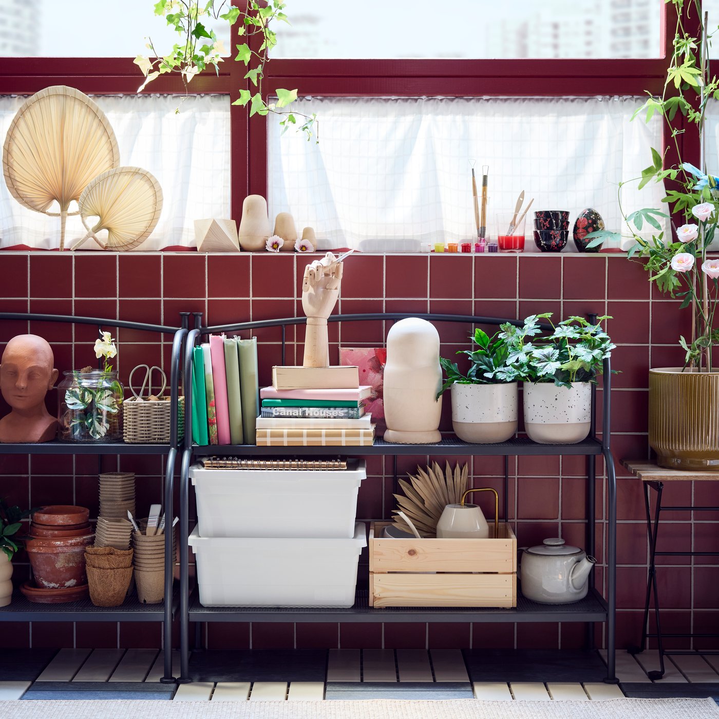 A bright enclosed balcony with two grey LÄCKÖ shelving units made for outdoor  use, displaying various plants and books.