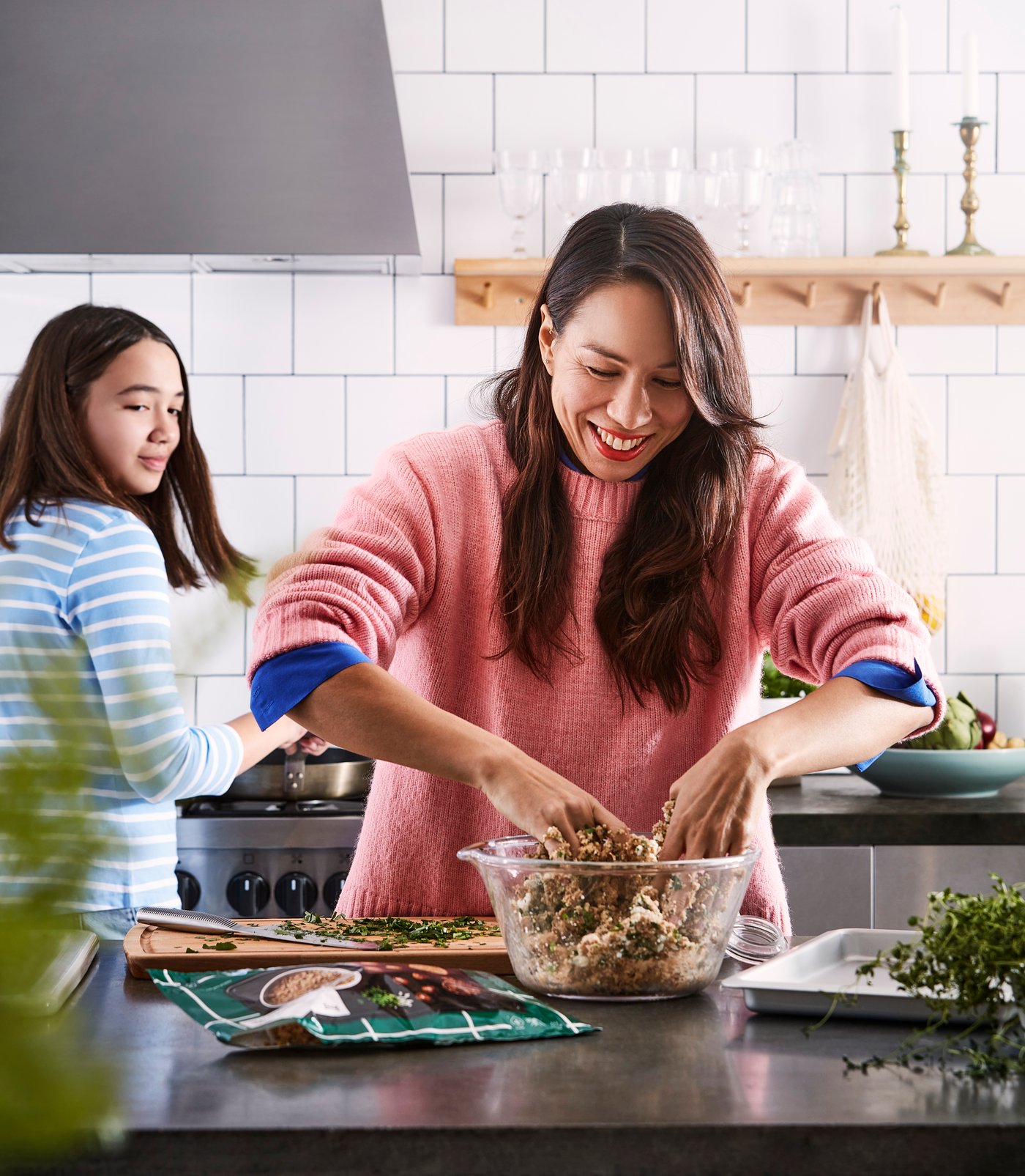 A person in a pink sweater prepares food in a kitchen, mixing ingredients in a bowl on the counter, while another person stands nearby at the stove.