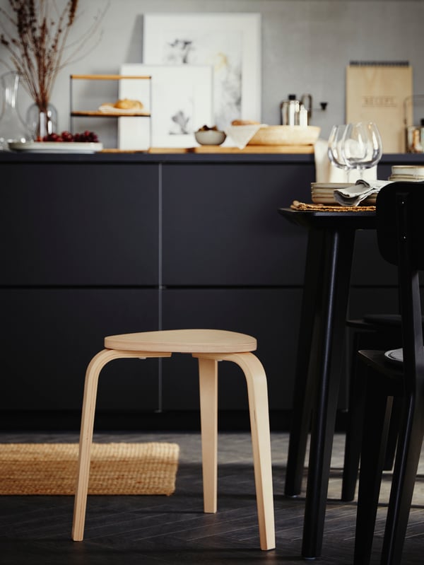 A three-legged stool in birch, a black cabinet with decorative items on the top surface and a table and two chairs in black.