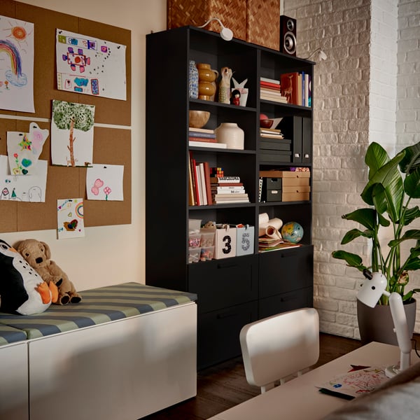 A black BRIMNES bookcase filled with books and decorative boxes, beside a cozy reading nook with soft-toy animals.
