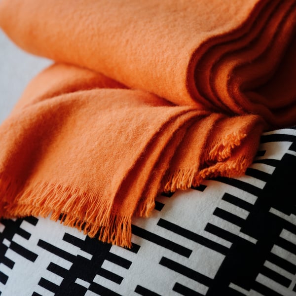 A close-up of a folded, bright orange STOCKHOLM 2025 throw resting on a cushion with a black and off-white cushion cover.