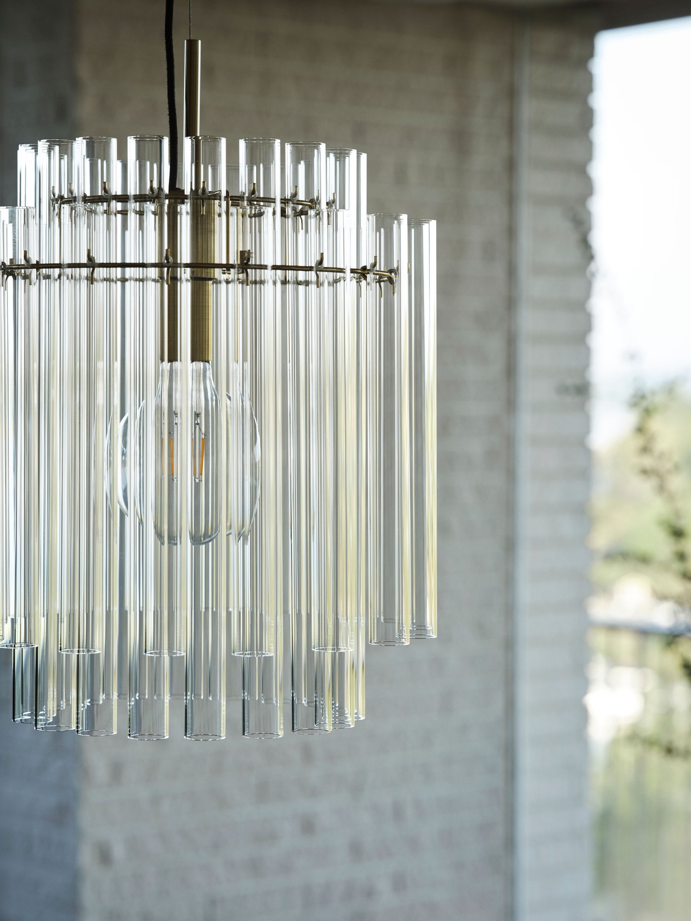 A brass-plated STOCKHOLM 2025 pendant lamp with glass tubes with a white brick wall in the background.