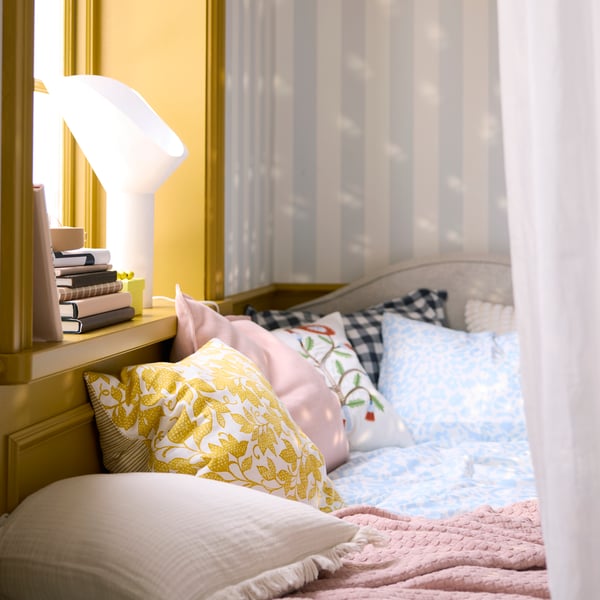 A close-up of the FJUNKLOCKA cushion cover in yellow and white, placed among various textured pillows on a cosy bed.
