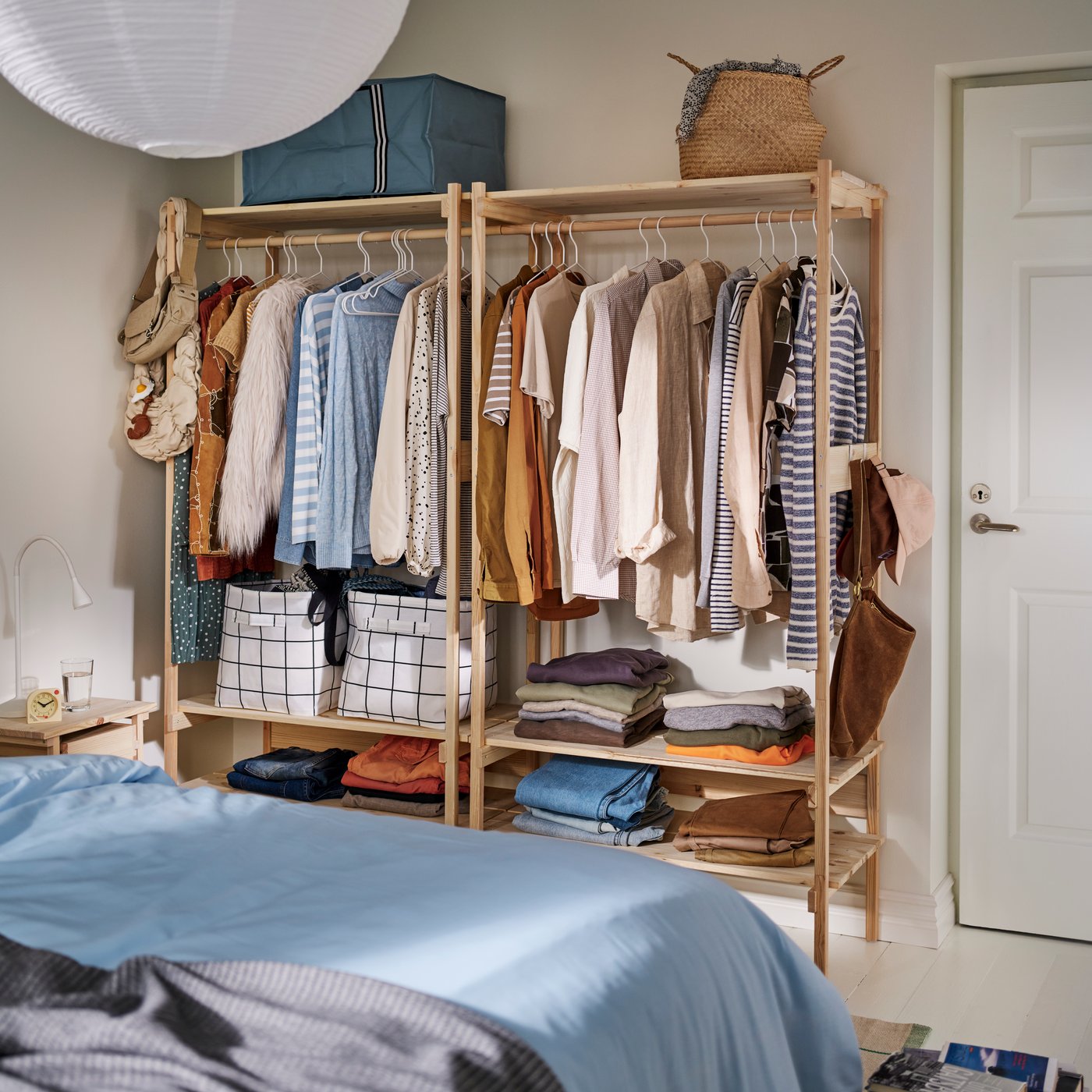 Two KRONÖREN open wardrobes in pine filled with hanging clothes, folded garments and storage baskets in a bedroom.