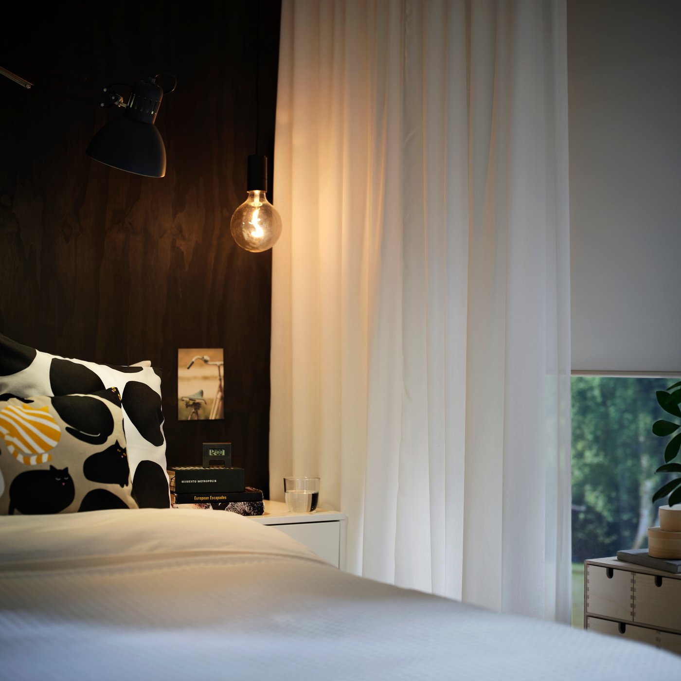 ROSENROBINIA sheer curtains in white hanging beside a bed with dark wall and bedside lamp in a softly lit bedroom.