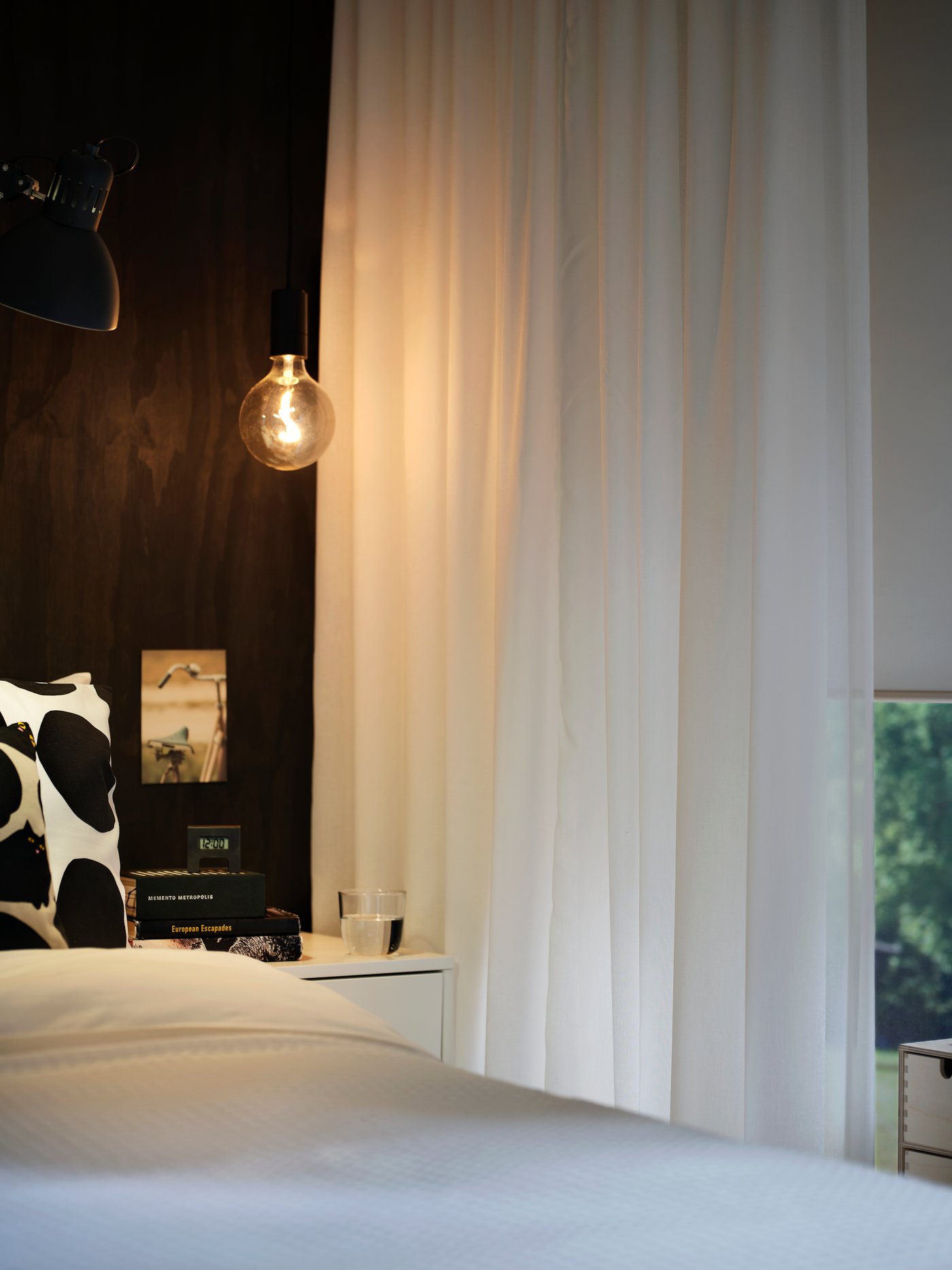 ROSENROBINIA‑Store in Weiß hängen neben einem Bett mit dunkler Wand und Nachttischlampe in einem sanft beleuchteten Schlafzimmer.