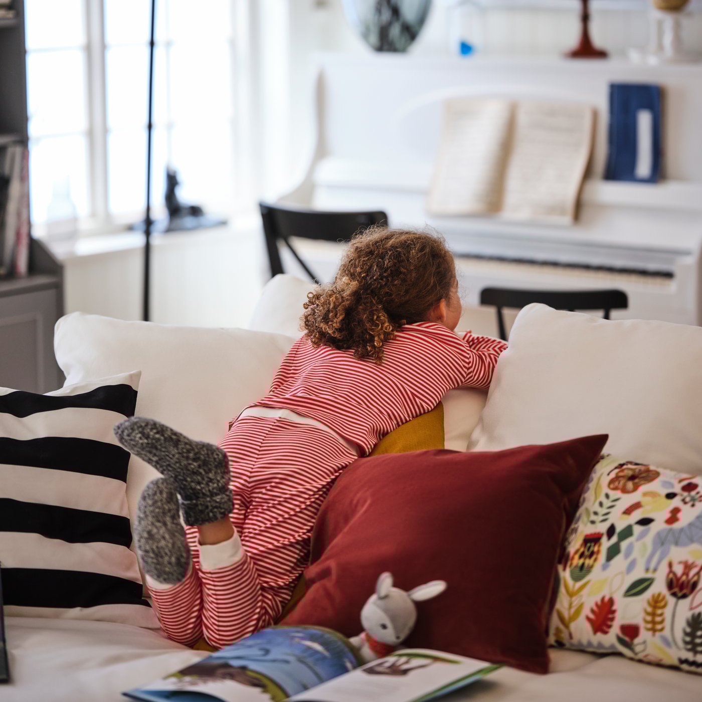 A young girl leans into the backrest of a white GRÖNLID four-seat sofa with chaise longue, peeking over its back.