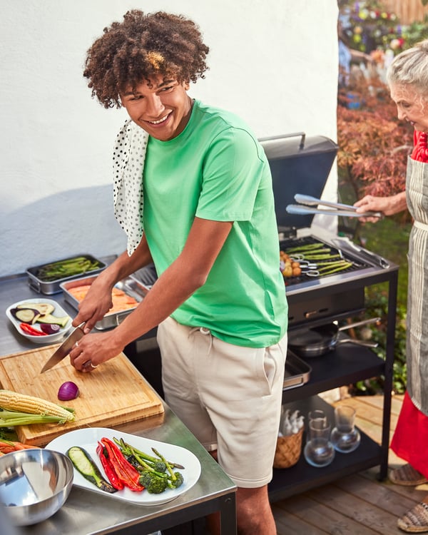 A boy in a green T-shirt is cutting vegetables on a wooden cutting board next to a GRILLSKÄR barbecue.