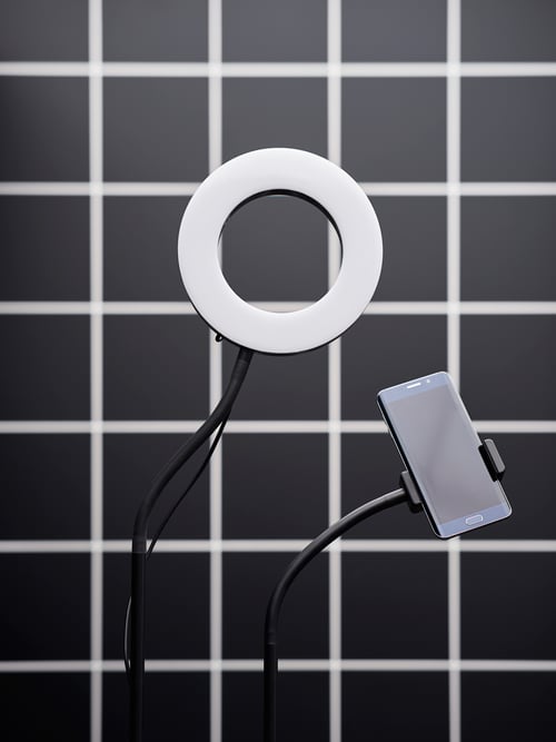 Nahaufnahme einer LÅNESPELARE Ringleuchte mit Mobiltelefonhalter vor einer schwarzweiß gefliesten Wand.
