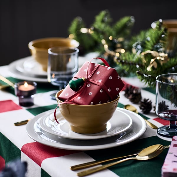 En liten present ligger i en skål på ett festligt dukat bord, inslagen i ett rött VINTERFINT-presentpapper med snöflingemönster.