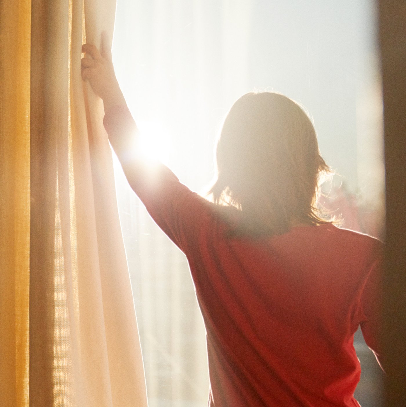 Dítě v červeném tričku roztahuje žluté a bílé závěsy, aby umožnilo slunečním paprskům proudit do místnosti.