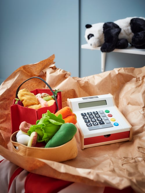 Camera copiilor cu o casă de marcat bej pentru jucării, un set bej de legume de jucărie, un set de coșuri de cumpărături pentru jucării, o jucărie moale panda.