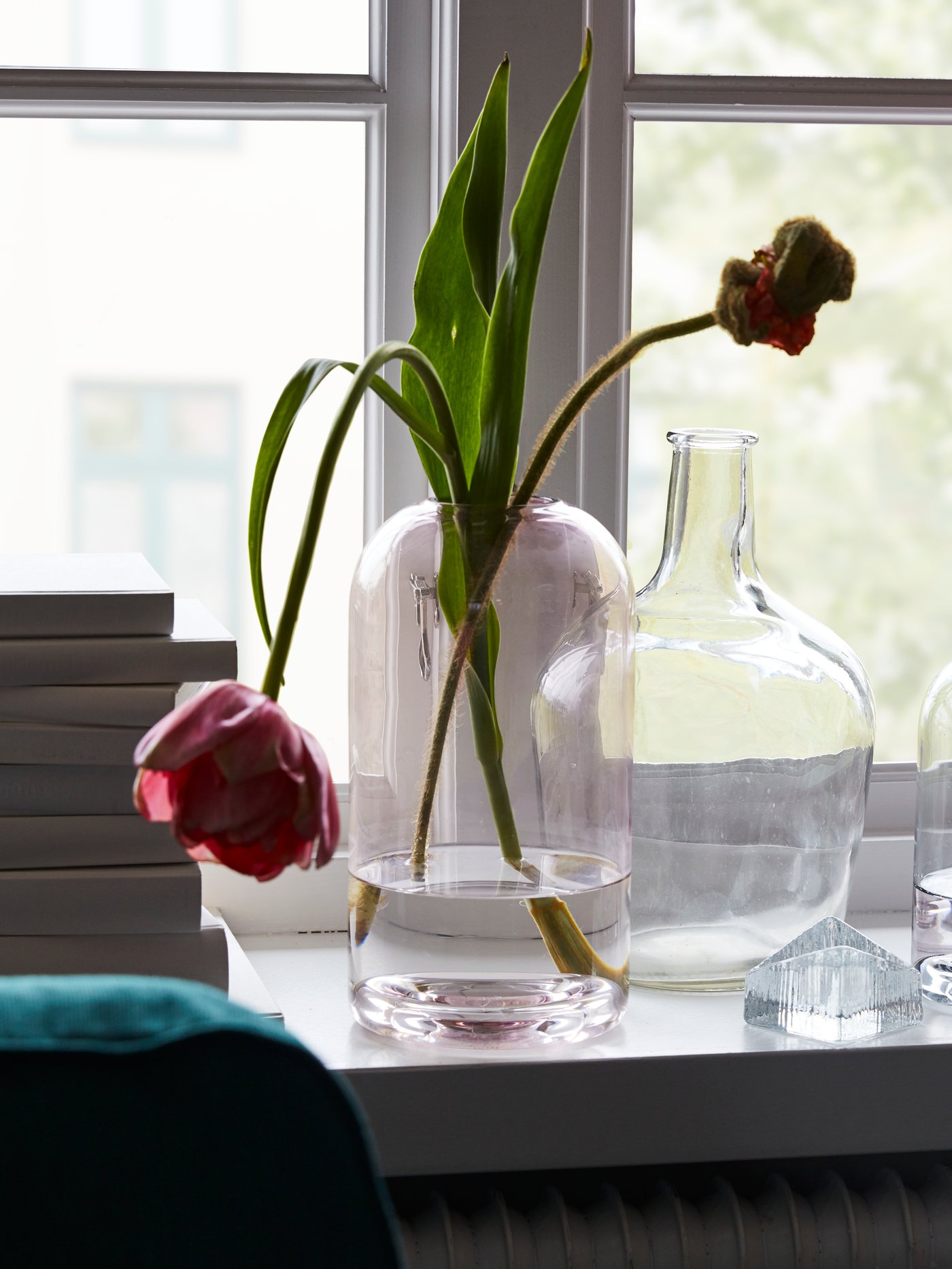 Clear and pink glass vases with flowers stand on a white window sill, the edge of a pile of books beside them.