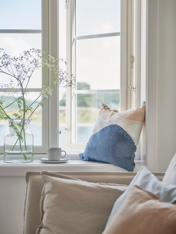 Fönsterbänk med ljus dyna och dekorativ kudde i blått, vitt och beige. På fönsterbrädan står en glasvas med vita blommor och en GLADELIG kopp på fat. Fönstret är öppet med utsikt över grönska och vatten.
