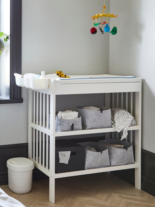 A white GULLIVER changing table with open felt boxes on its shelves and plastic racks of changing supplies hanging on an end.