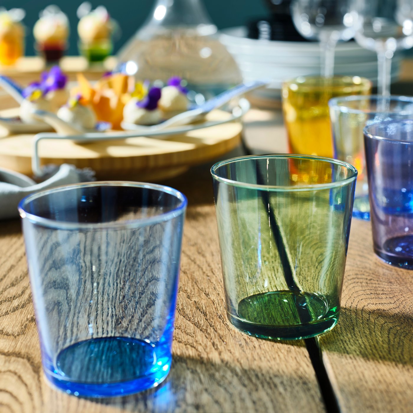 PAPPERSBJÖRK glasses, in blue, green and yellow, stand on a wooden table in front of a lazy susan with food.